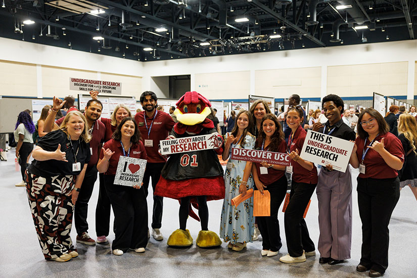 Staff and Undergraduate Research Ambassadors from the University of South Carolina Office of Undergraduate Research pose with Cocky at a Discover USC event, holding signs promoting undergraduate research.