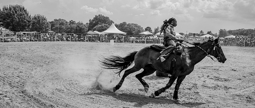 Black and white photograph of a young Black rider racing a horse around a dirt arena, dust rising behind them, with spectators and tents visible in the background under a wide Southern sky.