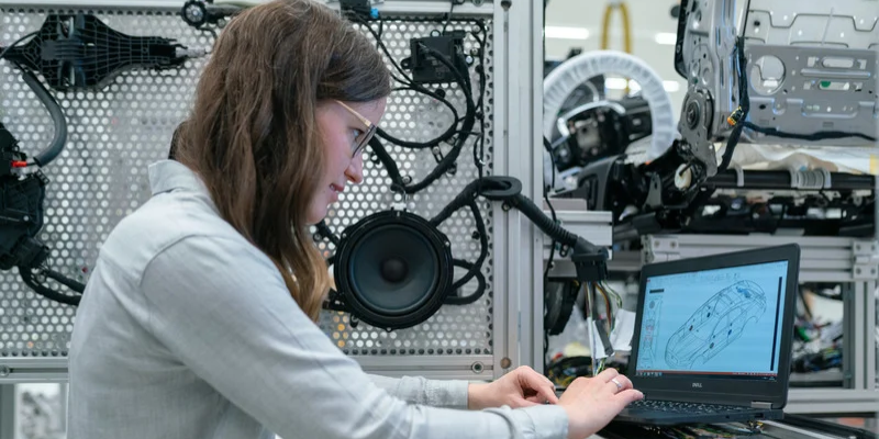 Person working on a laptop displaying a digital vehicle model, with automotive components in the background.