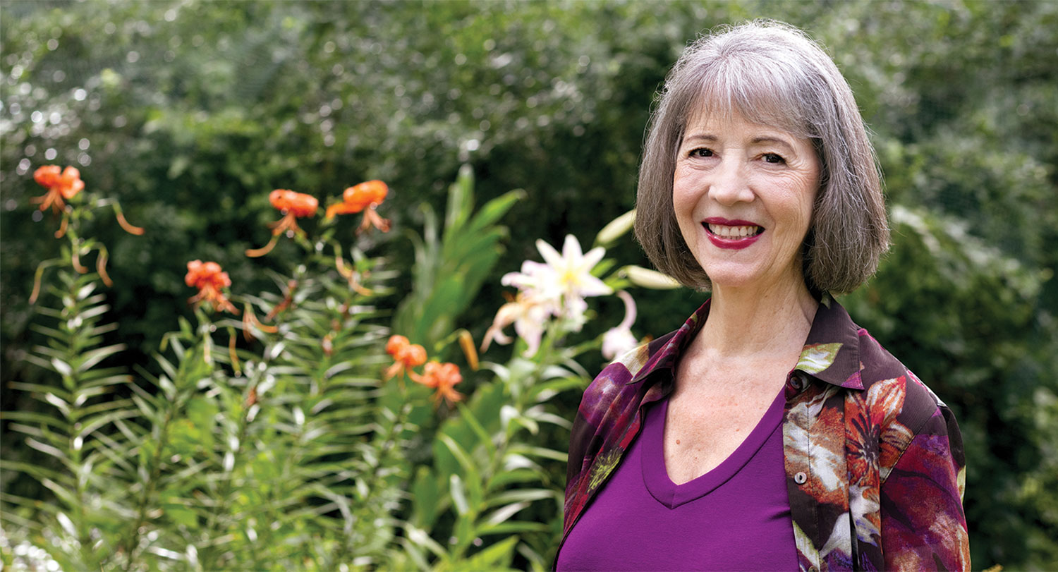 Retired theater professor Sarah Barker smiles for the camera with flowers in the background.
