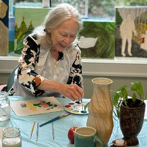 A woman paints a still life of jars and other items.
